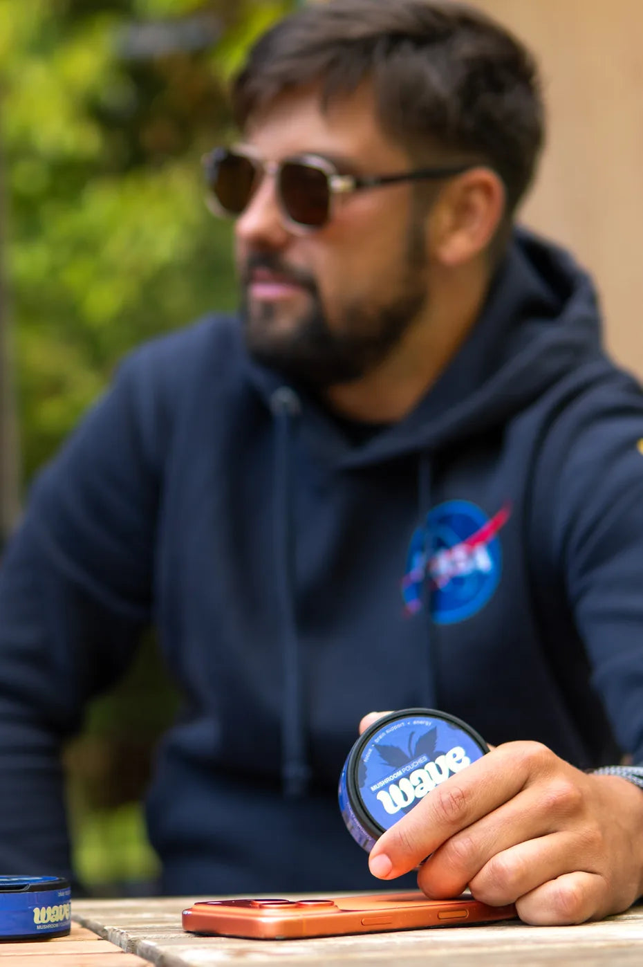 Mann mit Sonnenbrille und dunkelblauem Hoodie an einem Tisch im Freien, eine Dose WAVE in der Hand